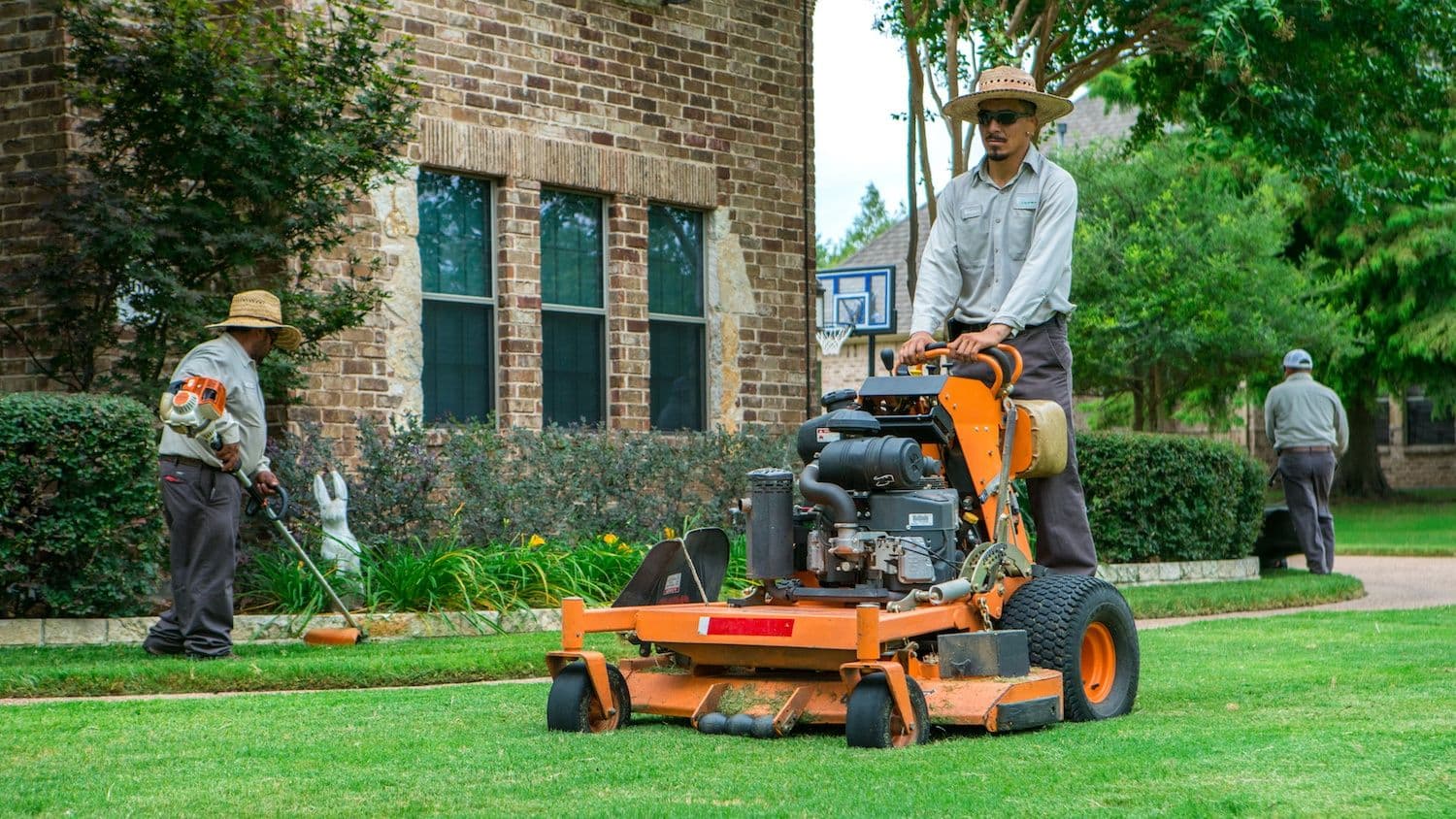 Landscapers maintain a residential lawn using a stand-on mower and a string trimmer.