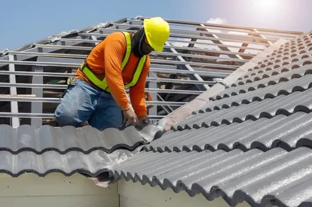 Construction worker in a yellow hard hat installs grey tiles on a metal roof frame.