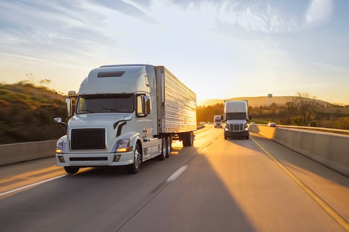 White semi-trucks driving on a highway during a golden sunset with long shadows and hills.